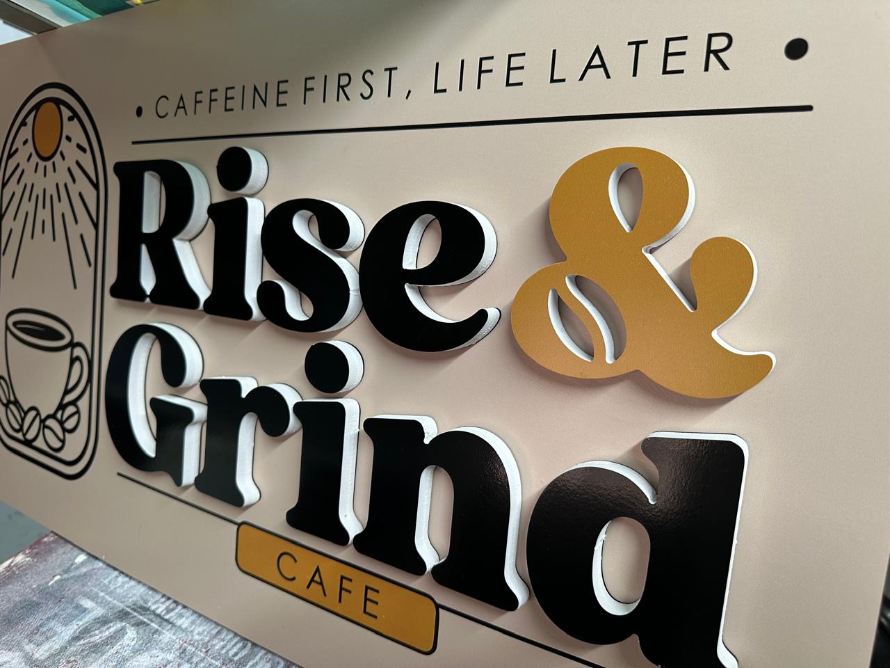 Rise & Grind café sign with black raised lettering, a yellow ampersand and coffee illustration.
