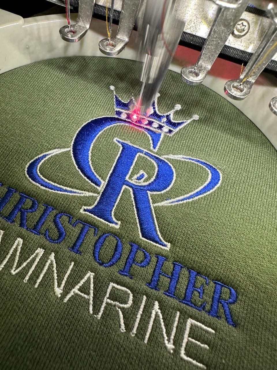Embroidery machine stitching a blue and white crowned CR design on olive fabric.