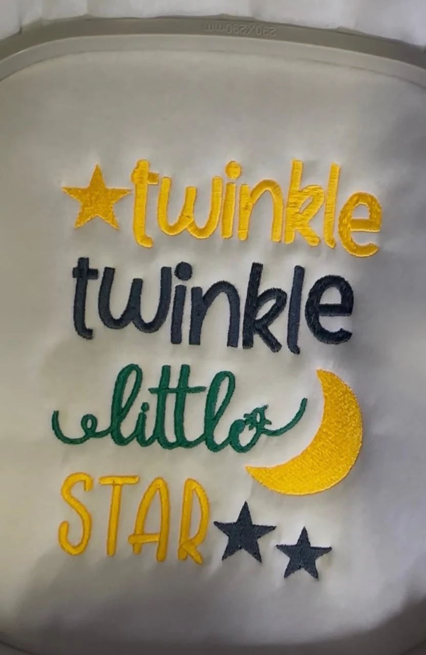 White fabric in an embroidery hoop with multicoloured Twinkle Twinkle Little Star lettering, stars and a crescent moon.